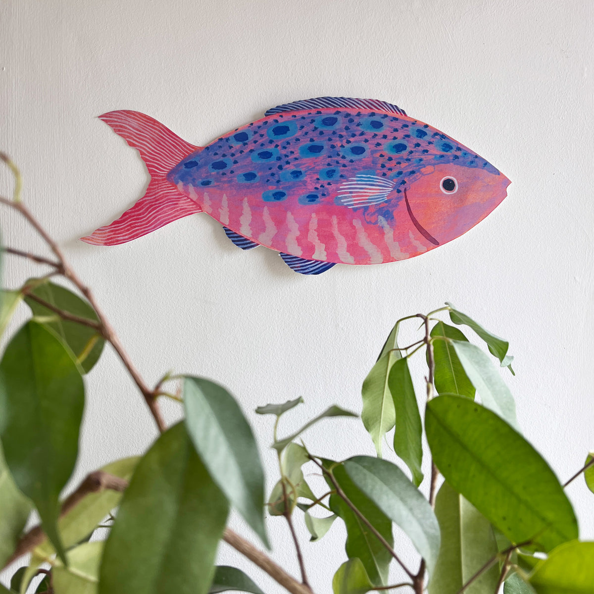 Large wooden fish decoration by Lee Foster-Wilson displayed on a white wall with a green plant in the foreground