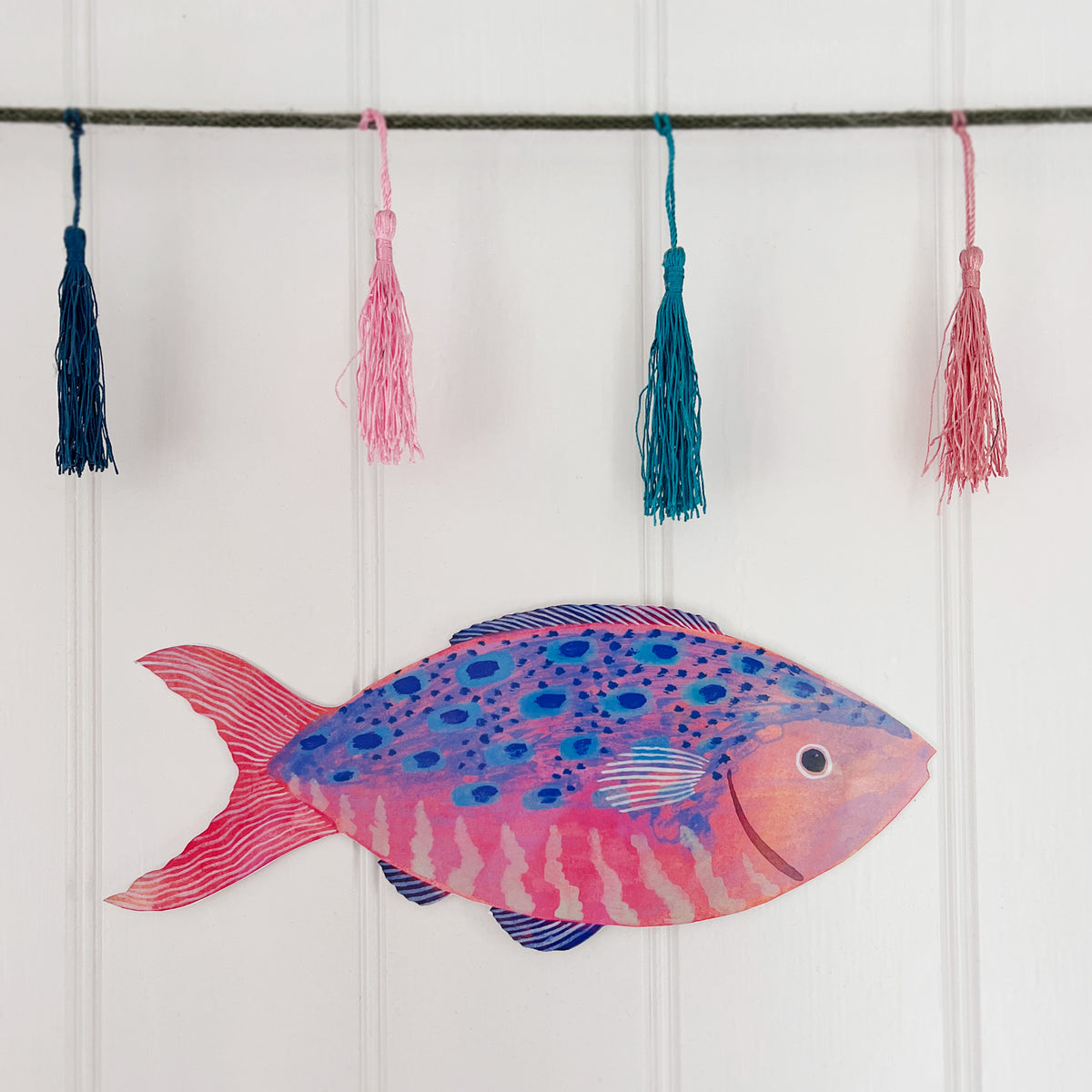 Large wooden fish decoration by Lee Foster-Wilson displayed on a white tongue and groove wall with colourful tassels above