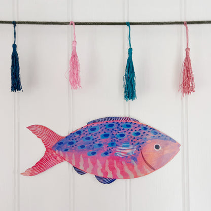 Large wooden fish decoration by Lee Foster-Wilson displayed on a white tongue and groove wall with colourful tassels above