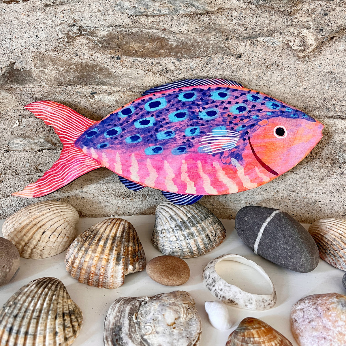 Large wooden fish decoration by Lee Foster-Wilson displayed on real shells against a stone wall