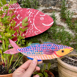 Colourful mackerel fish decoration by Lee Foster-Wilson being held up with plants and a giant painted fish in the background