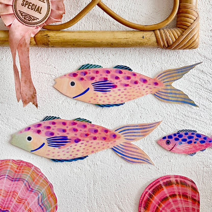 Colourful fish decorations by Lee Foster-Wilson displayed on a rough textured wall beneath a rattan frame